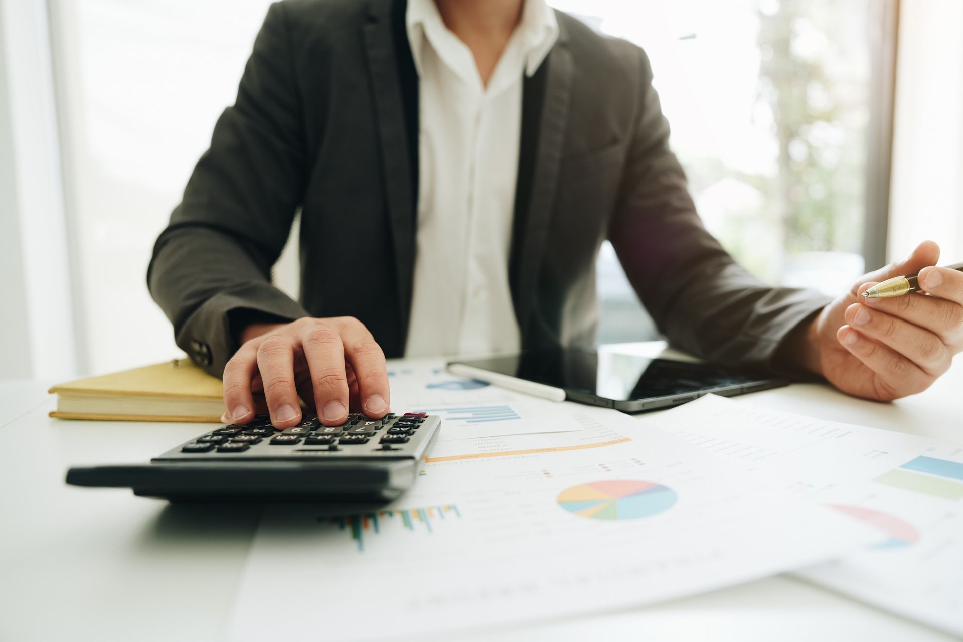 Una persona vestida de traje utiliza una calculadora en un escritorio junto a documentos financieros y un bolígrafo.