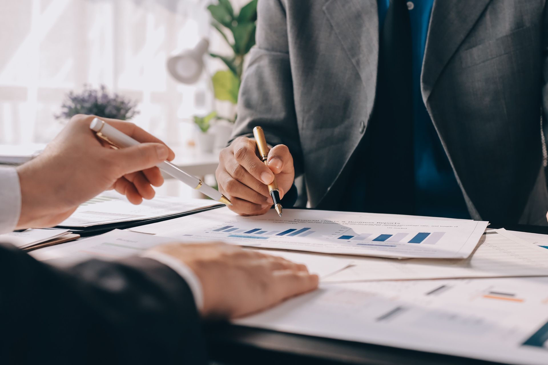 Dos personas vestidas con traje revisan gráficos y documentos en un escritorio; una de ellas escribe.