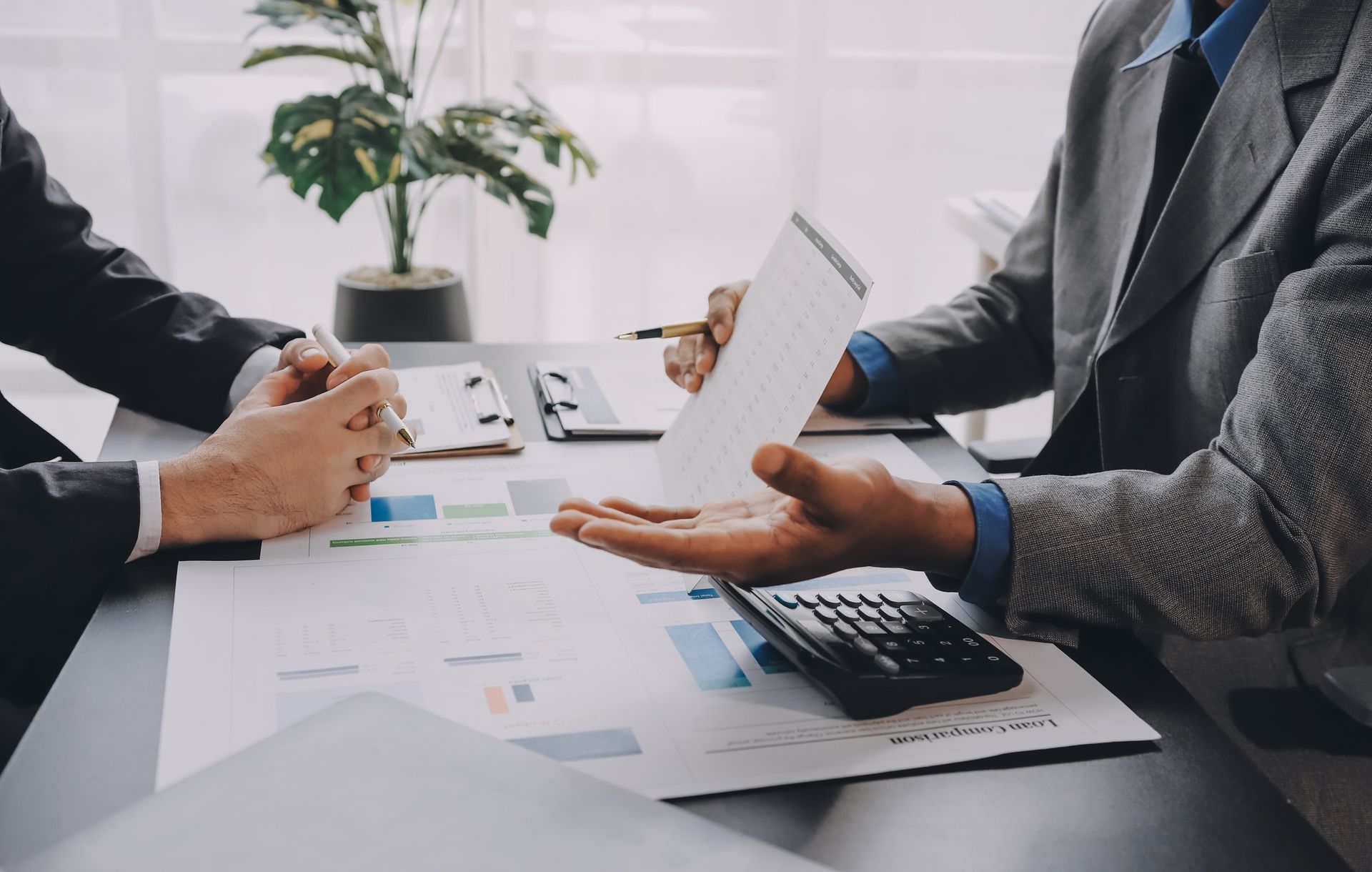 Dos personas con traje, sentadas a una mesa, revisan documentos, con una calculadora y una planta.