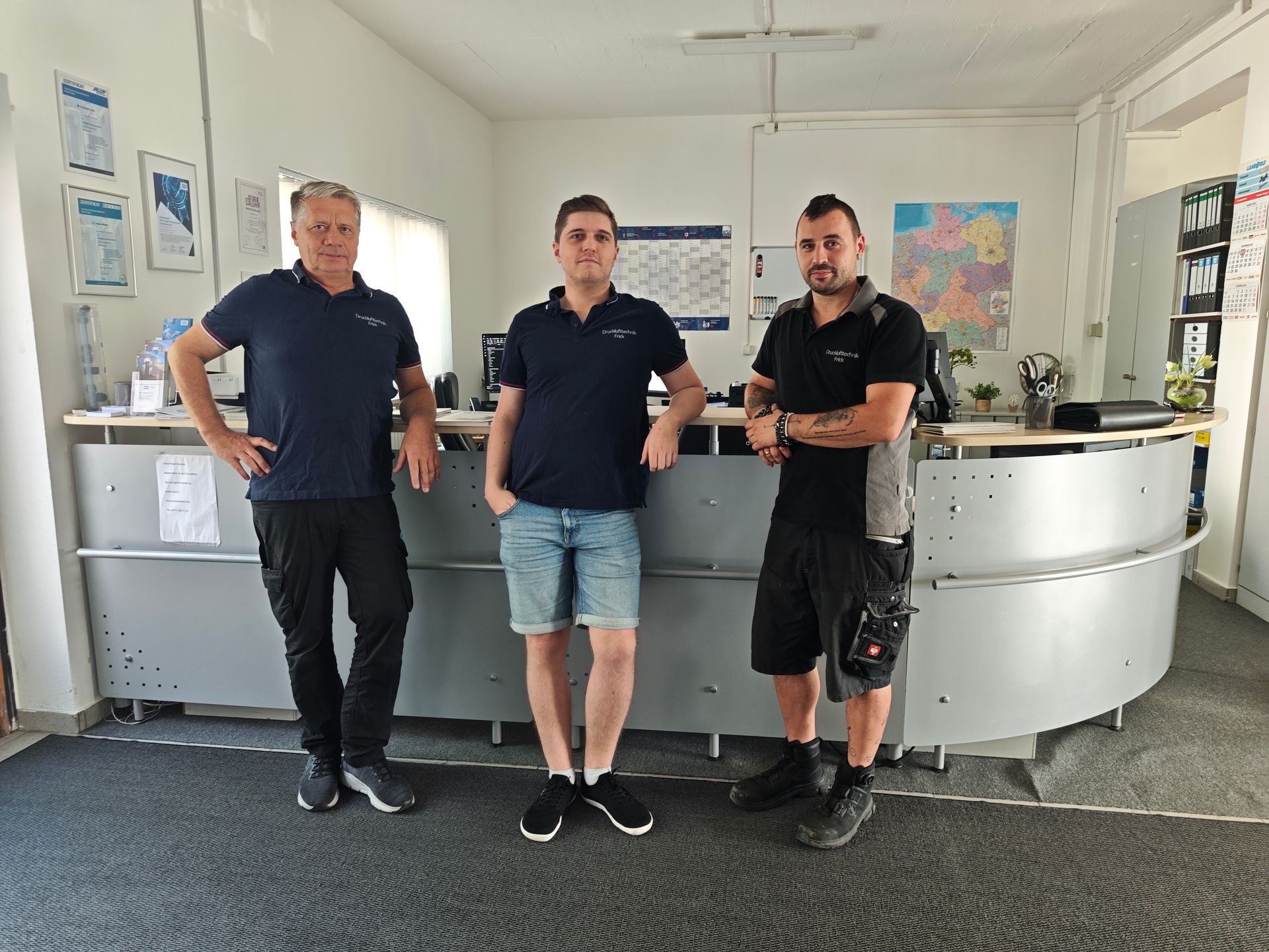 Drei Männer stehen hinter einem geschwungenen Empfangstresen; an der Wand hängen eine Landkarte und ein Kalender.