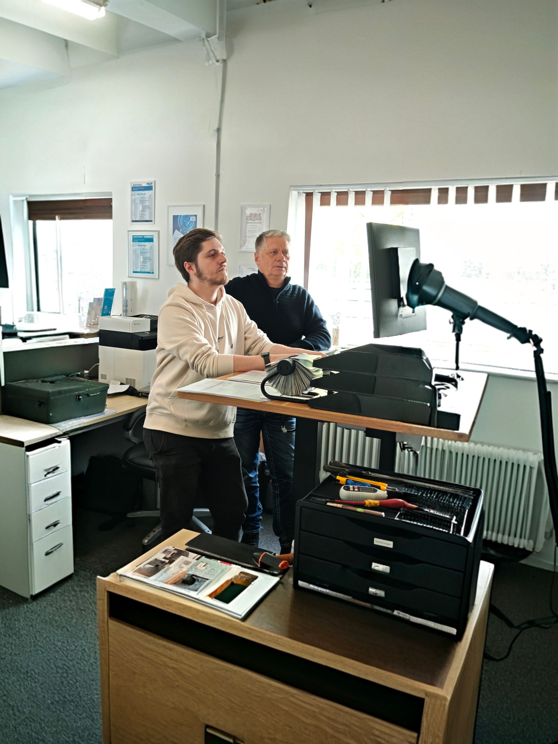 Zwei Männer stehen an einem Schreibtisch mit Geräten. 
