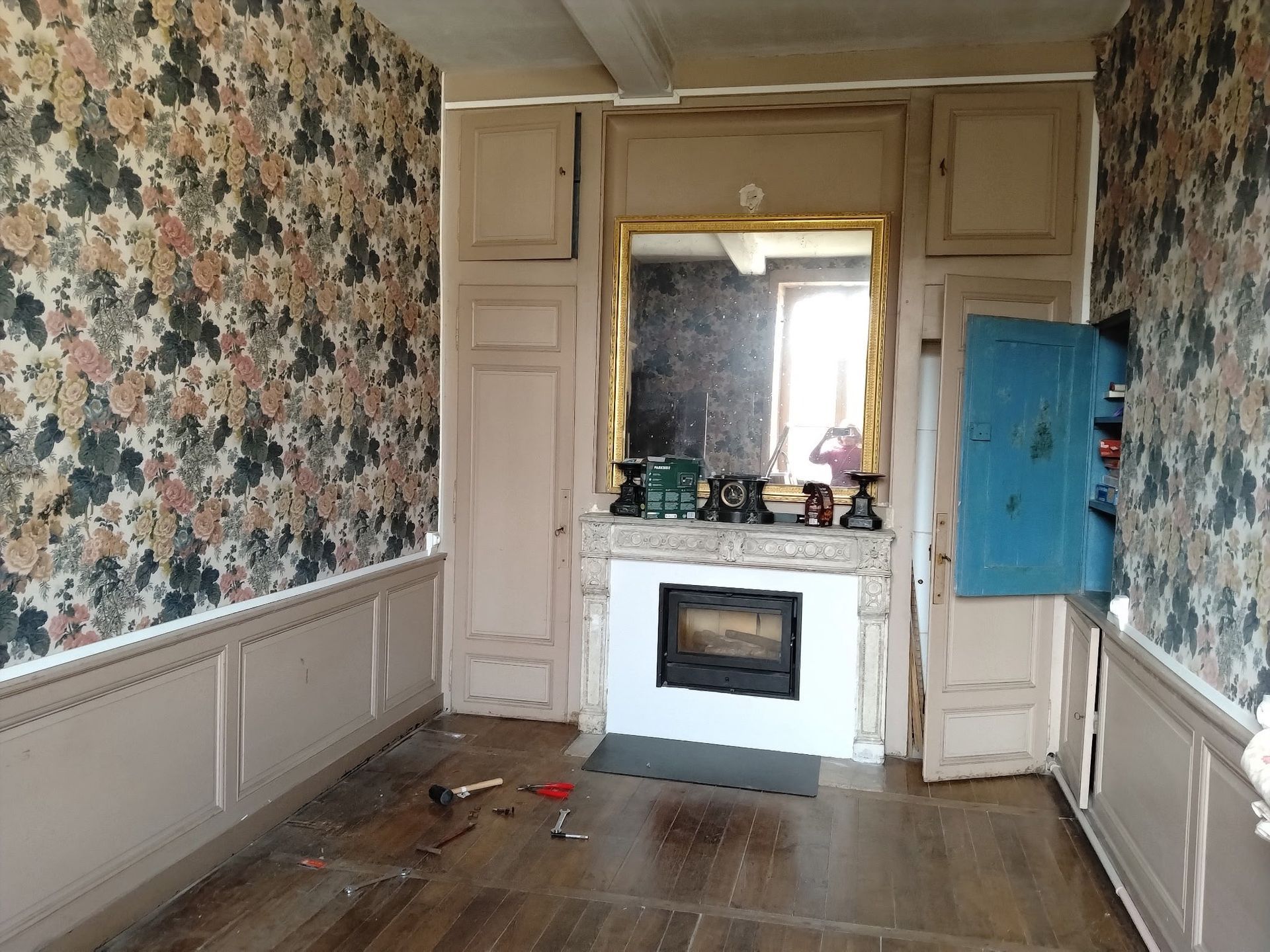 Pièce vide avec papier peint à motifs floraux, lambris blancs, cheminée et parquet.