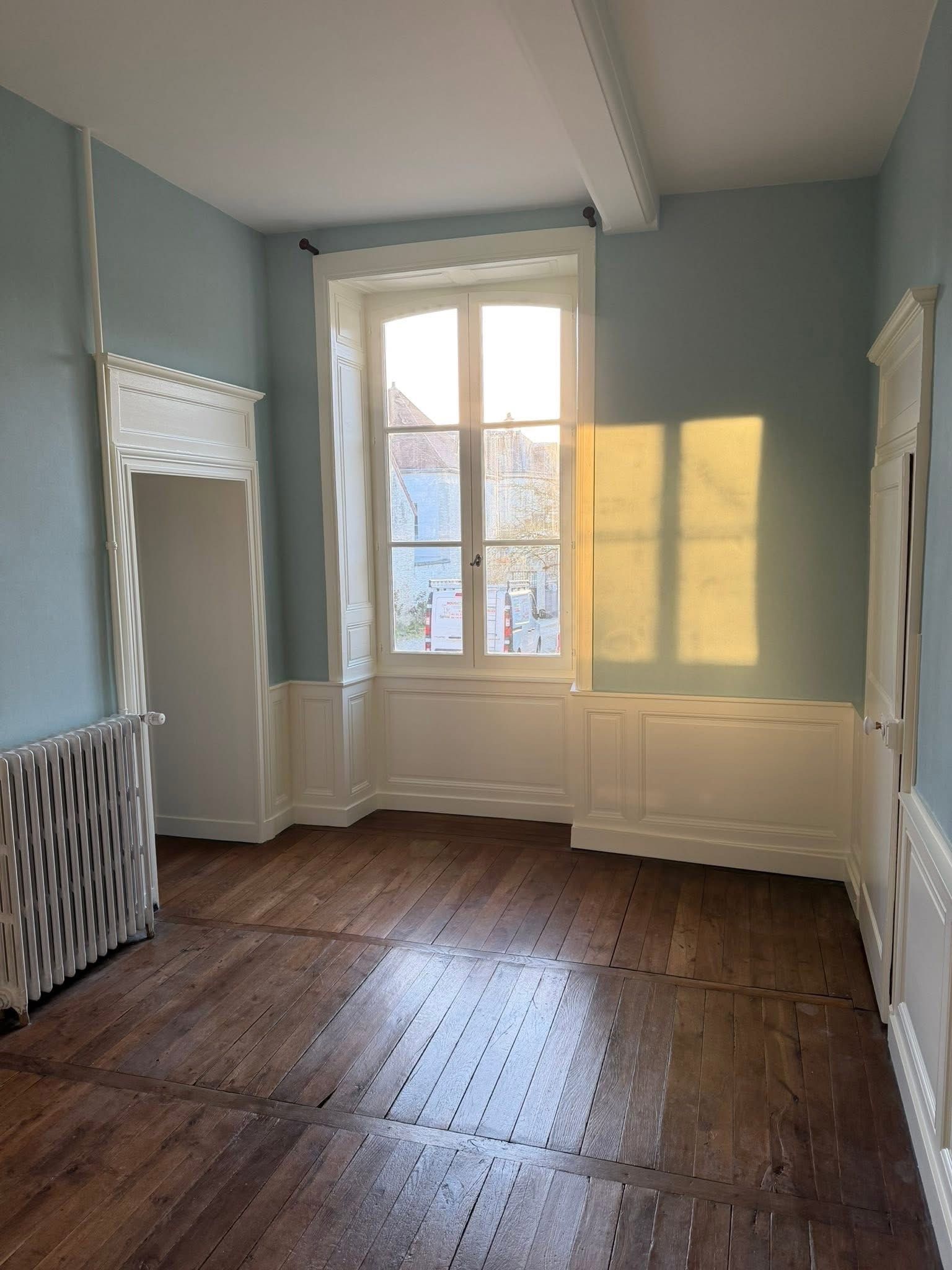 Pièce bleue vide avec parquet, haute fenêtre et radiateur sous la fenêtre.