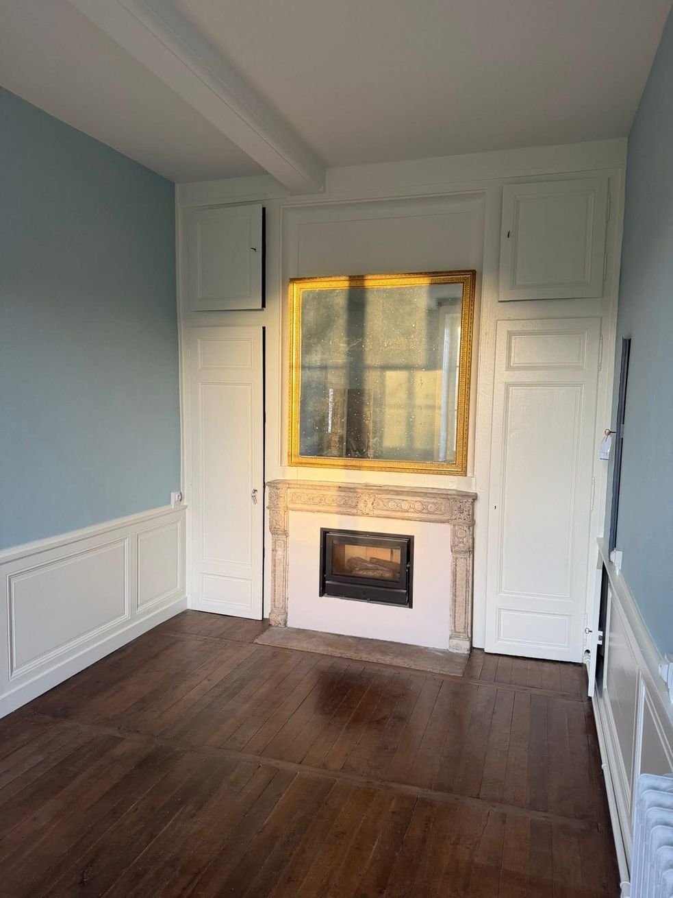 Pièce vide avec parquet foncé, murs blancs et cheminée encastrée sous un miroir à cadre doré.