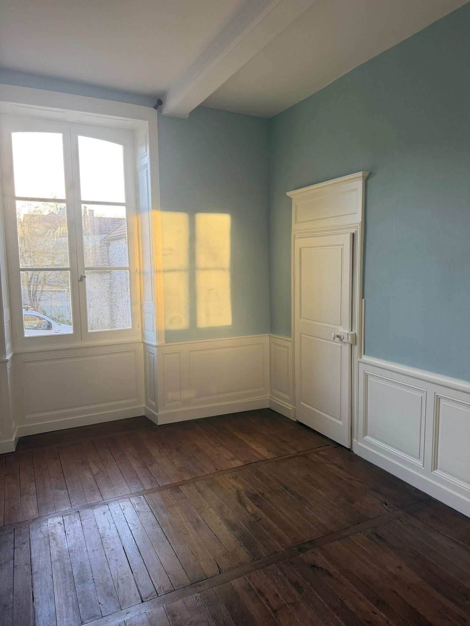 Pièce vide aux murs bleus, moulures blanches, haute fenêtre et parquet usé laissant entrer la lumière du soleil.
