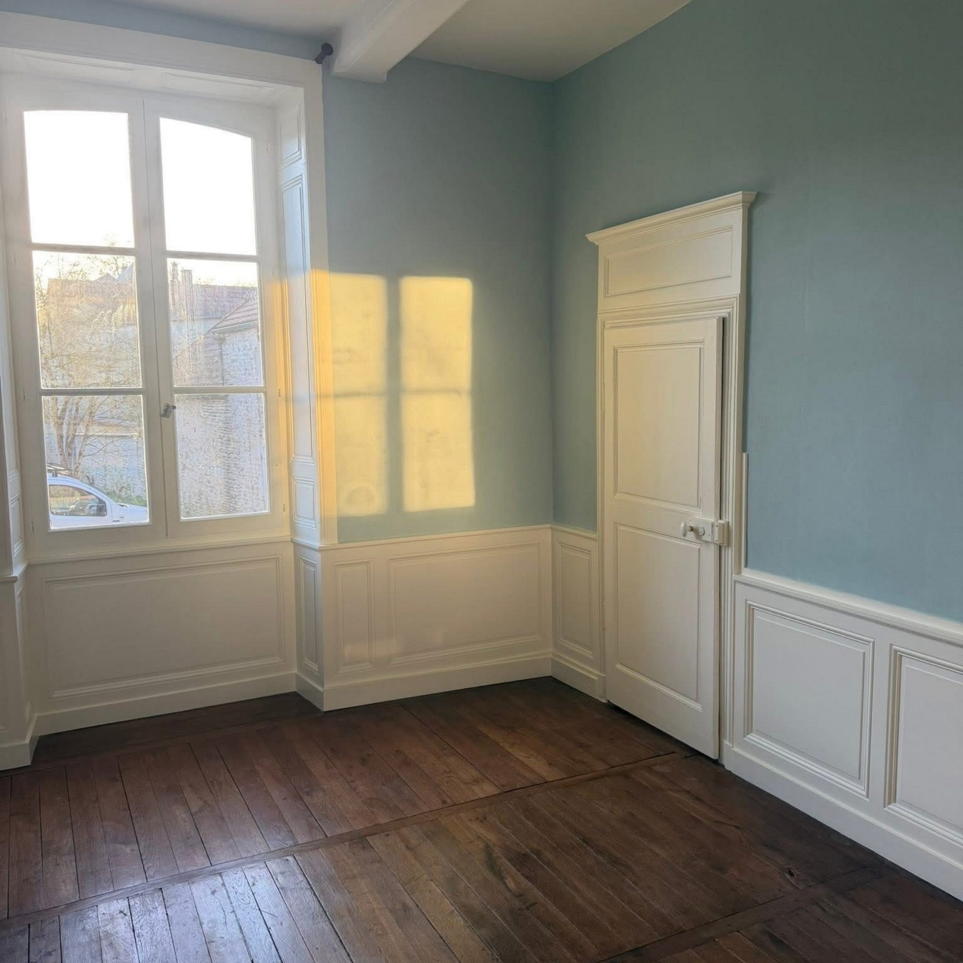 Pièce vide et ensoleillée aux murs bleus, aux moulures blanches, au parquet et avec une porte à côté d'une fenêtre.