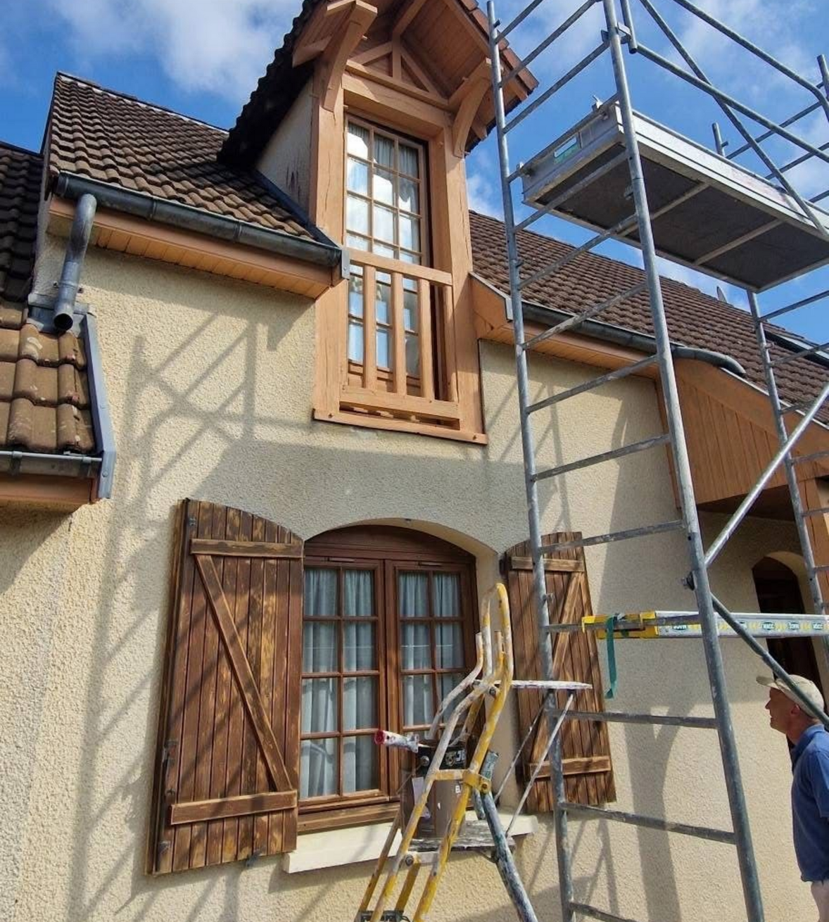 Façade d'une maison en rénovation, avec échafaudages et échelle, laissant apparaître des volets en bois ouverts et une lucarne.