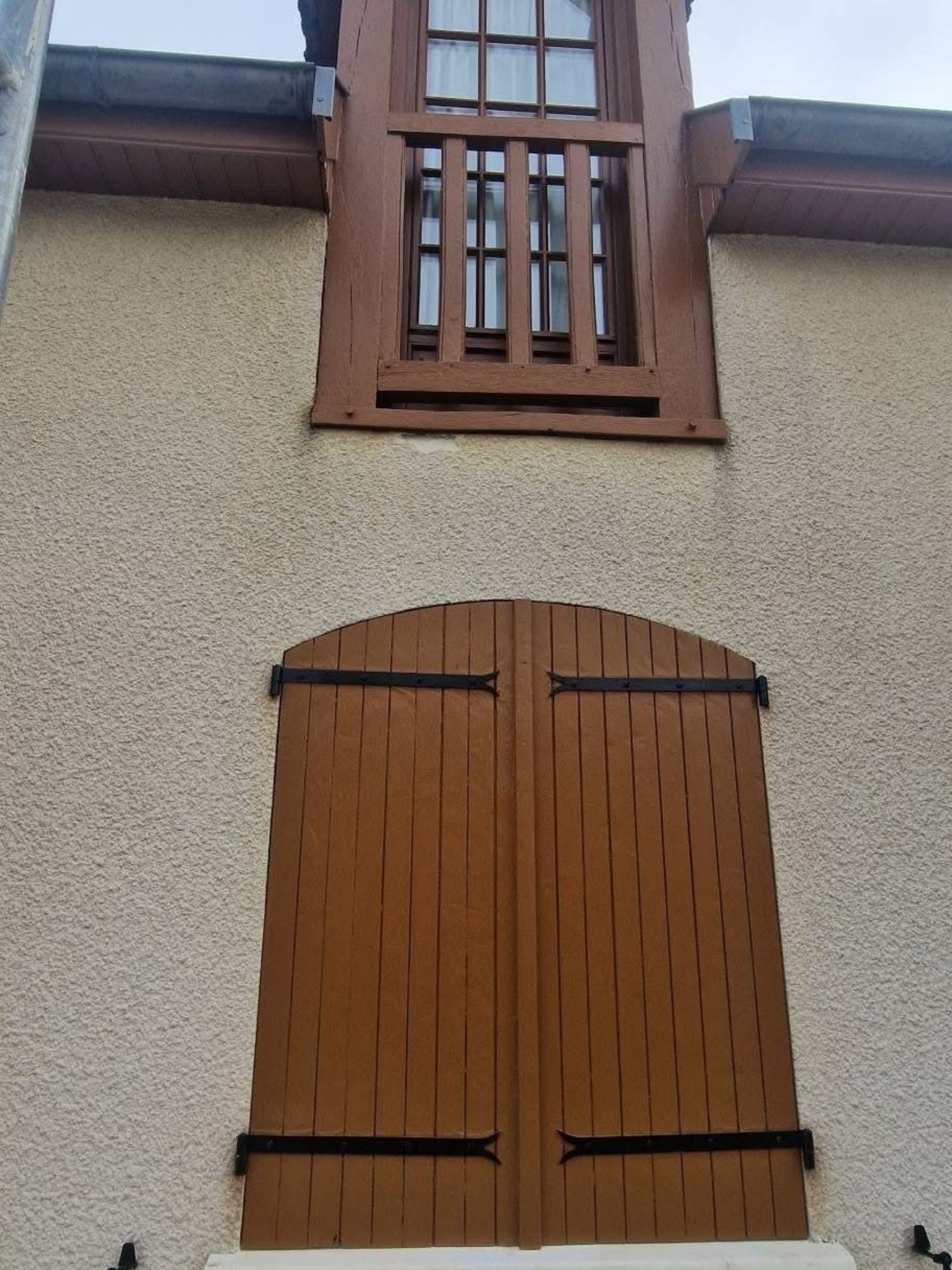 Mur en stuc beige avec une fenêtre à volets en bois brun au-dessus d'une porte cintrée en bois assortie.