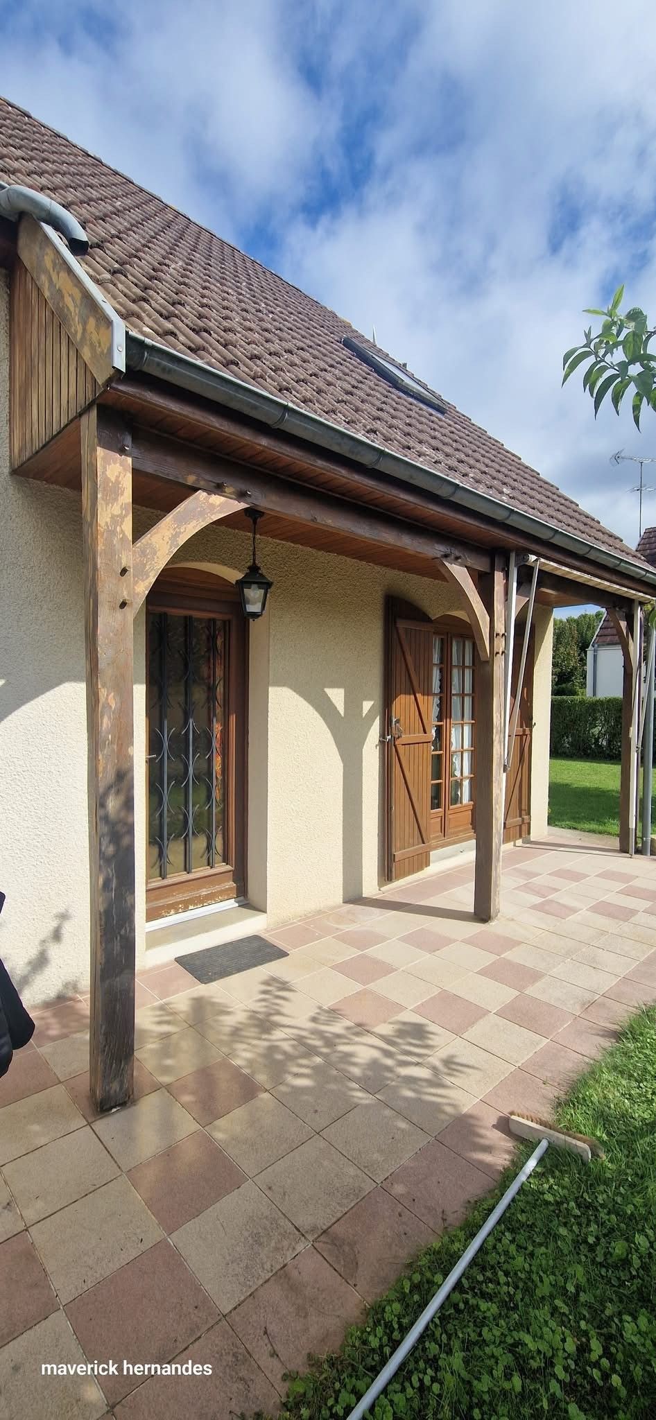 Porche d'une maison en briques avec poteaux en bois, porte cintrée et toit de tuiles, donnant sur un chemin de jardin.