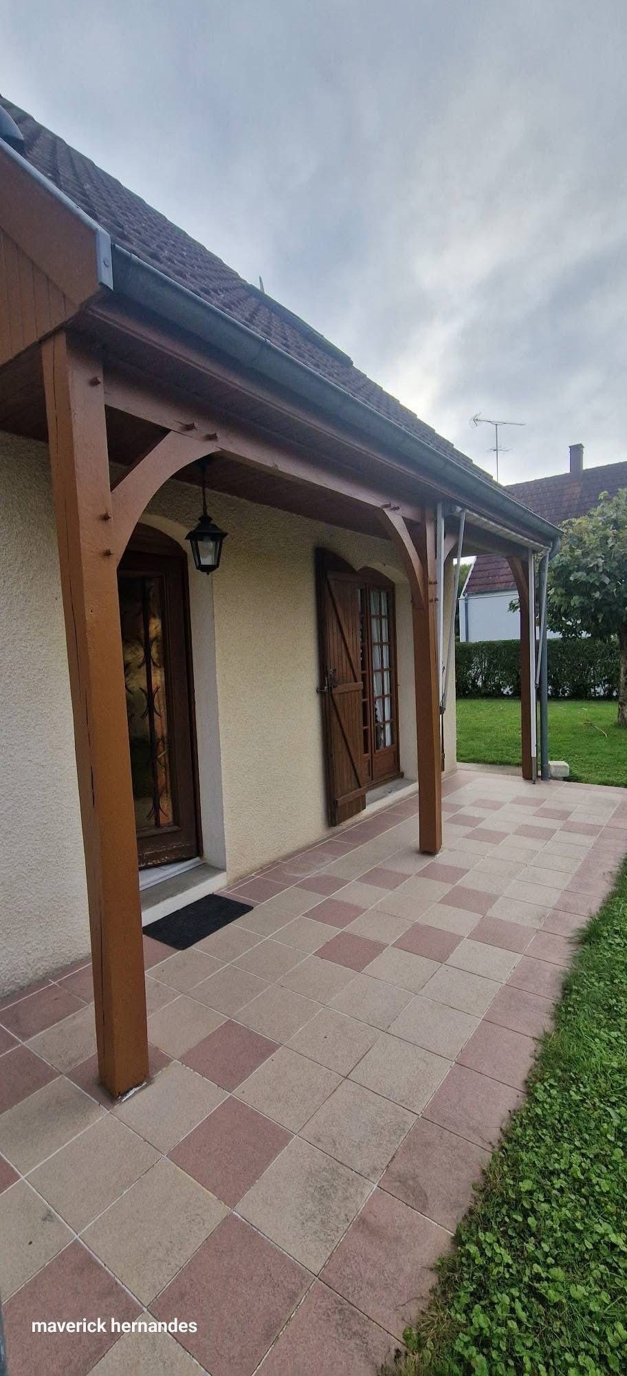 Maison en briques avec porche couvert, colonnes en bois, toit de tuiles et allée de jardin sous un ciel bleu.