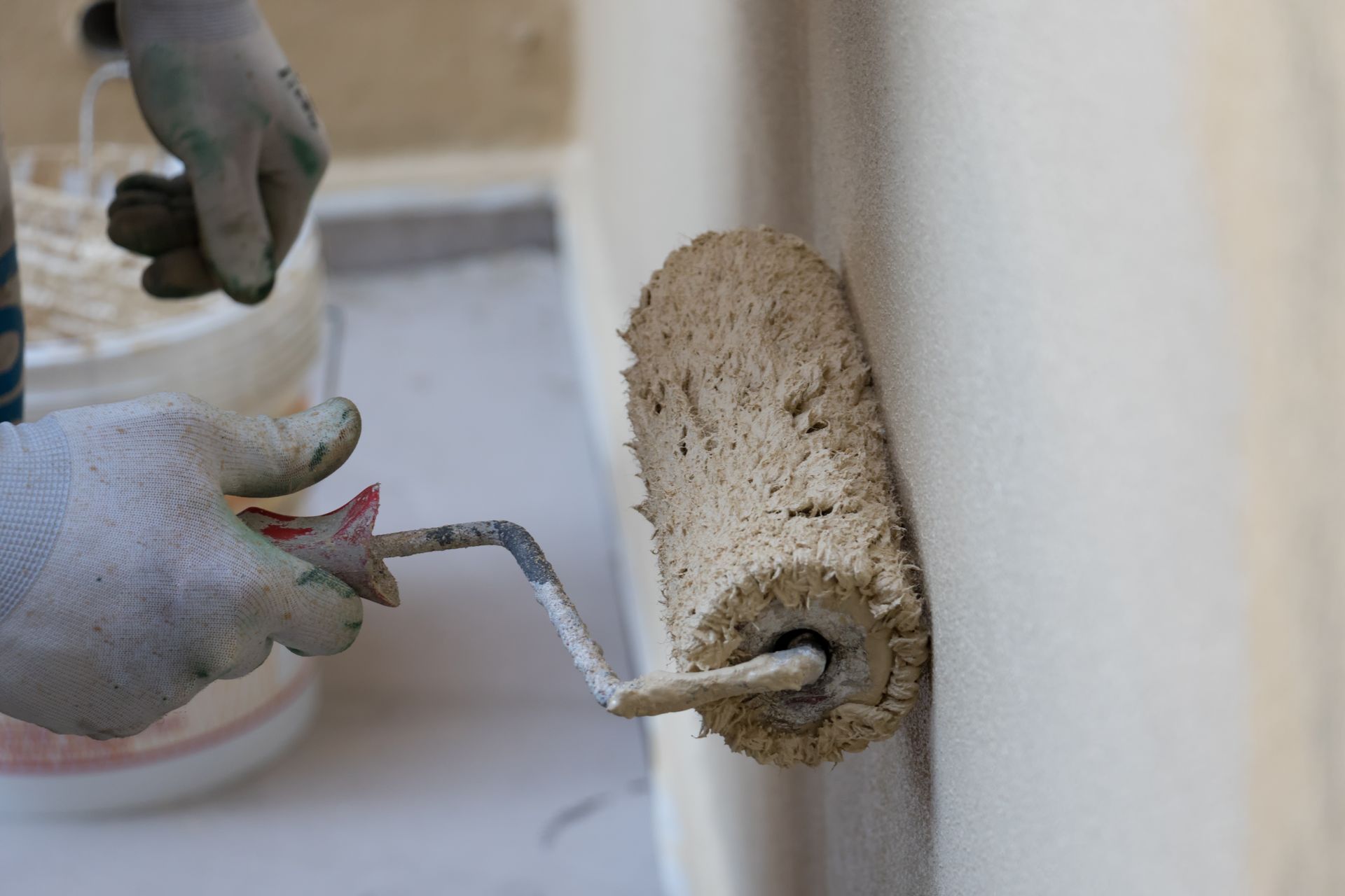 Des mains gantées utilisent un rouleau à peinture pour enduire un mur extérieur beige de peinture texturée.