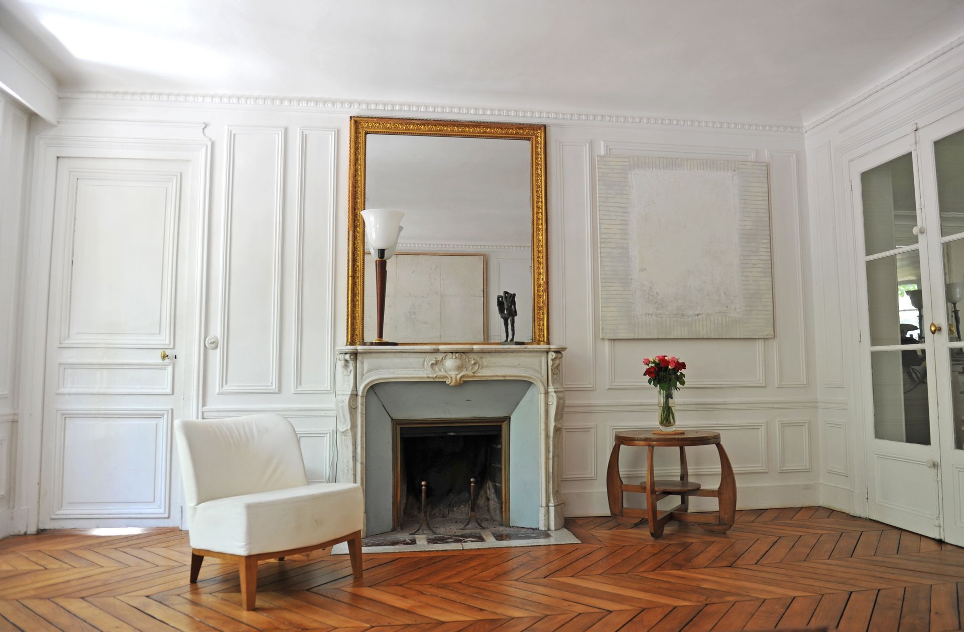 Élégant salon blanc avec cheminée, miroir doré, fauteuil, petite table et parquet.