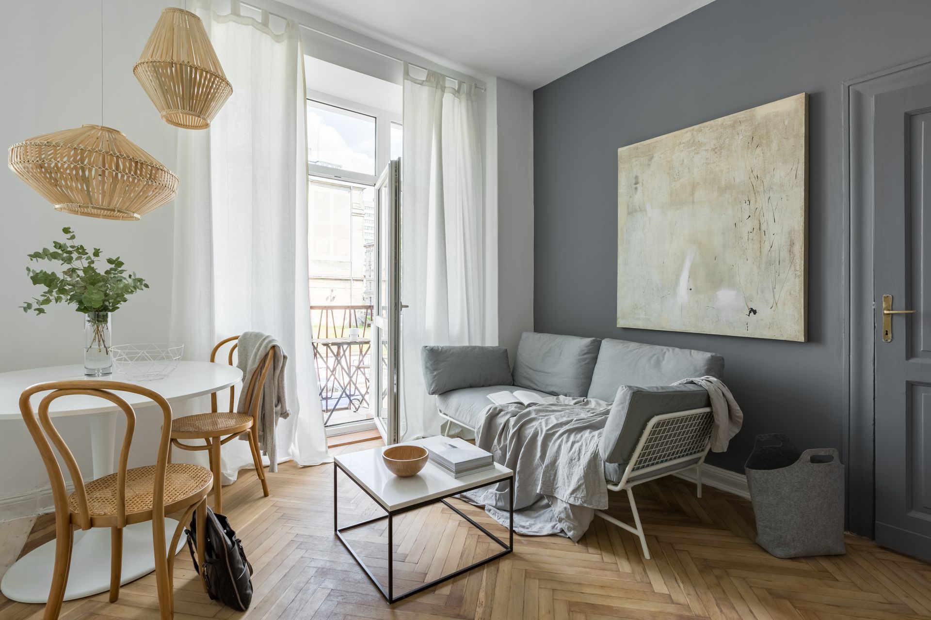 Salon moderne et lumineux avec canapé gris, fauteuils en osier, table à manger et grande œuvre d'art abstraite au mur.