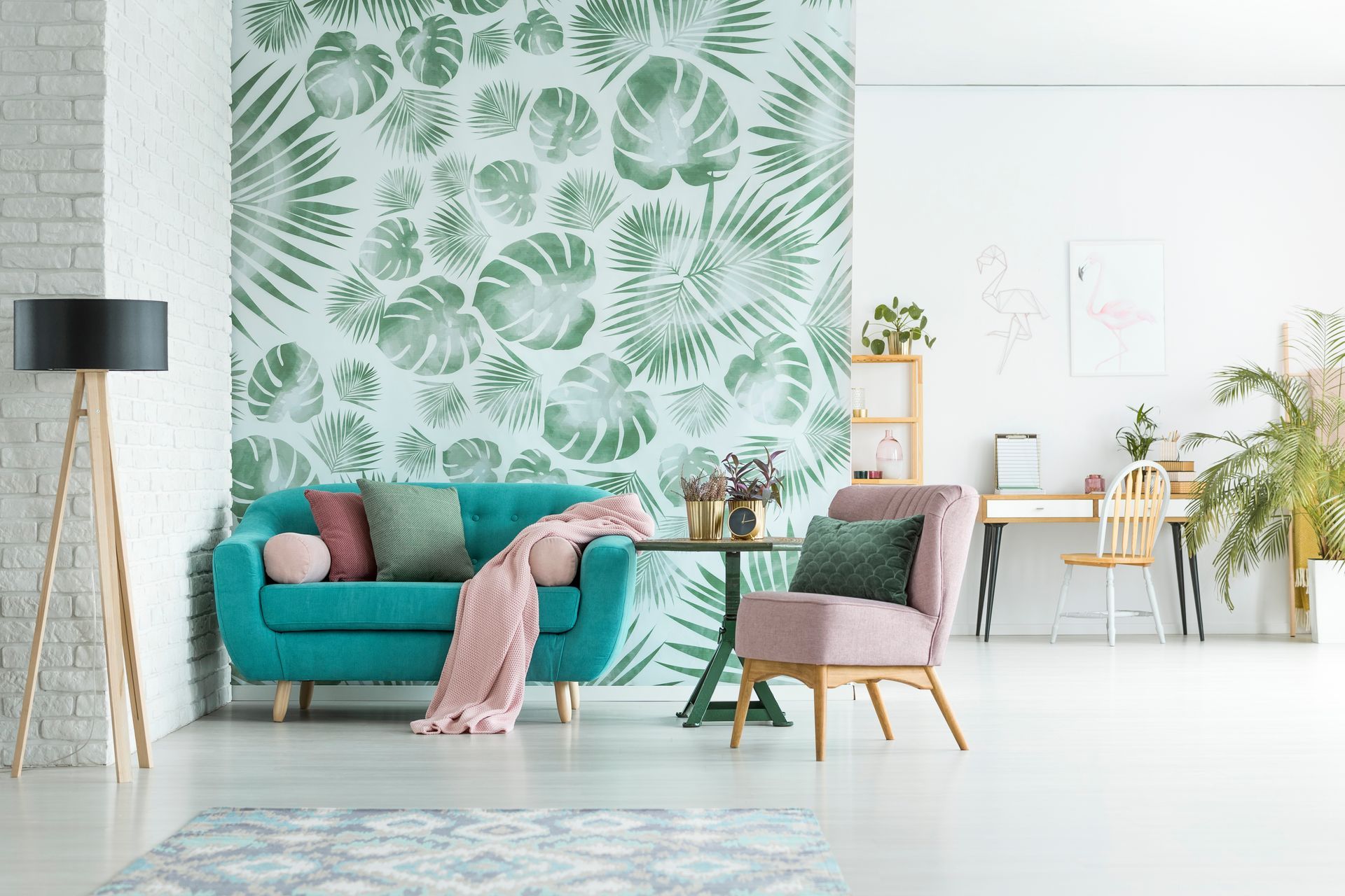 Salon lumineux avec canapé turquoise, plaid rose, fresque murale à motifs de feuilles tropicales et plantes près d'un bureau.
