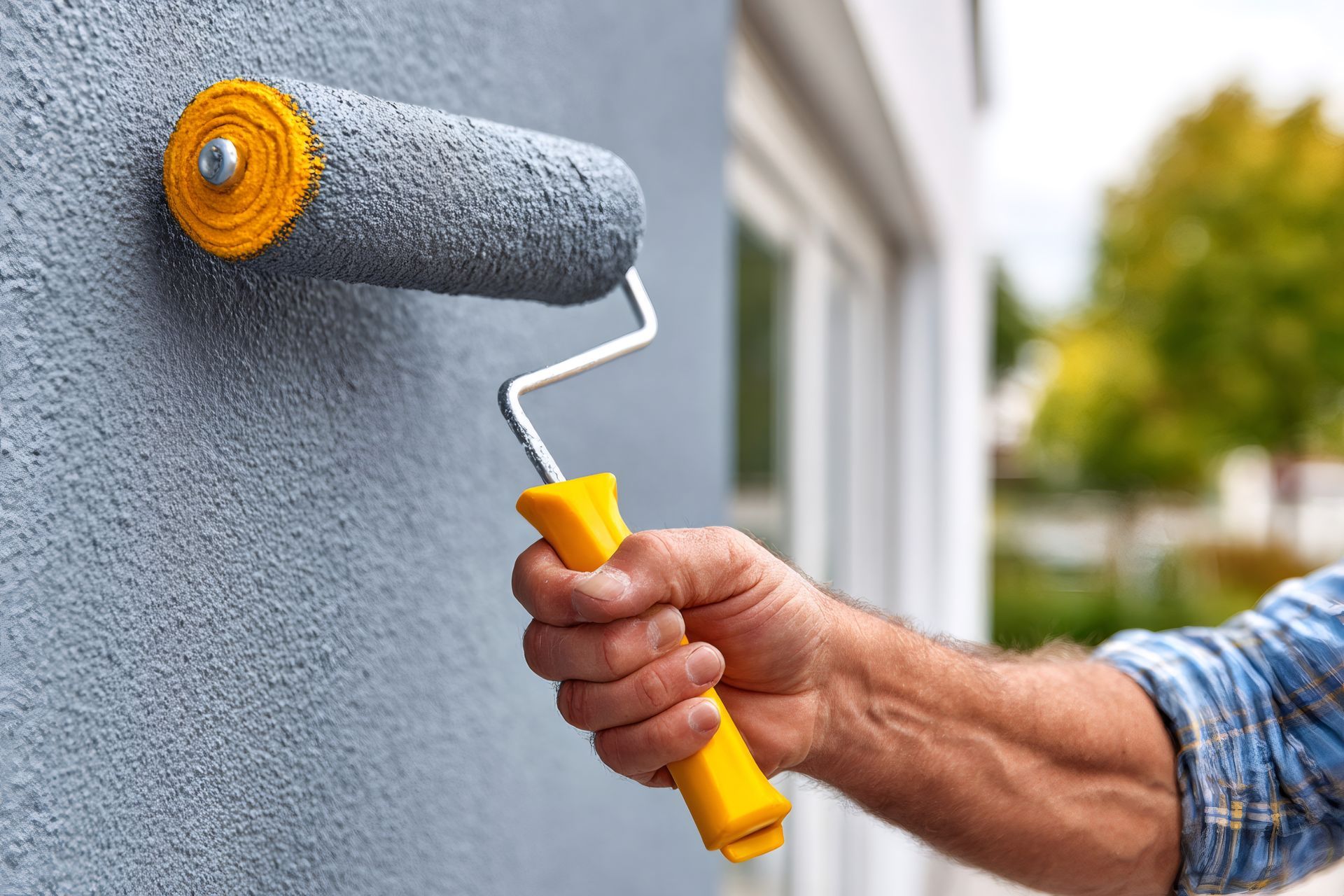 Une personne peint un mur gris à l'extérieur avec un rouleau à peinture à manche jaune.