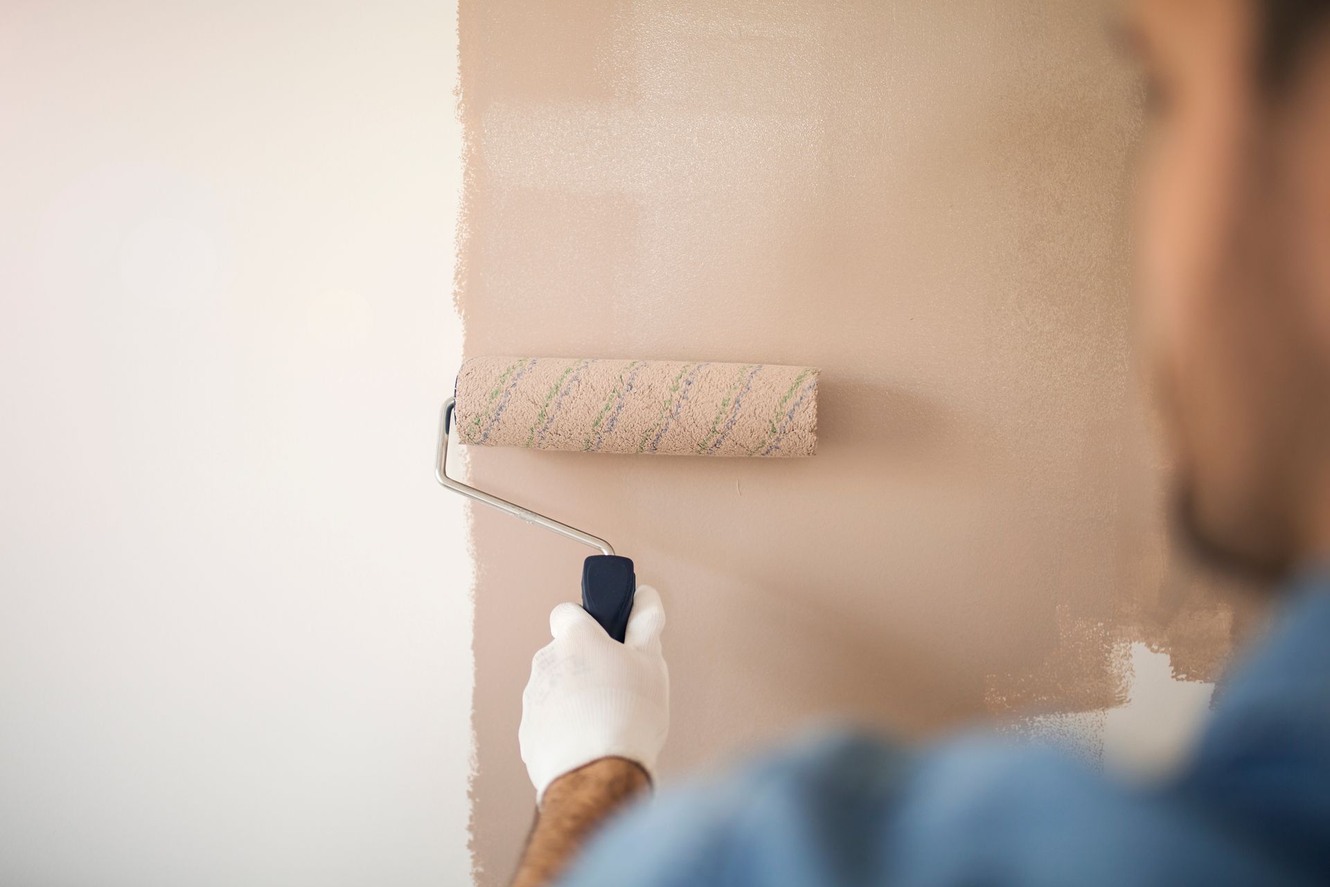 Une personne peignant un mur beige au rouleau, vue de près par derrière.