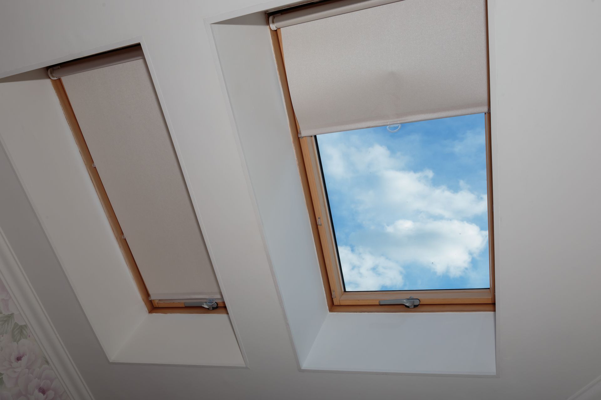 Deux fenêtres de toit dans un plafond blanc, l'une ouverte sur un ciel bleu nuageux.