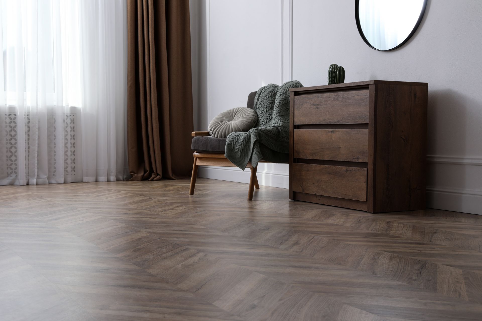Chambre vide avec une commode en bois, un fauteuil, un miroir rond et des voilages près d'une fenêtre.