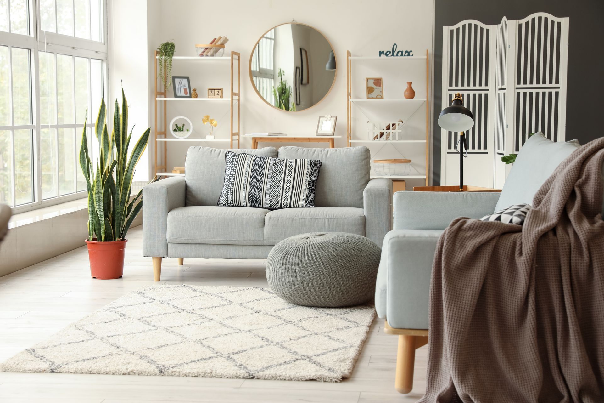 Salon lumineux avec canapé gris, miroir rond, étagères blanches, plantes et tapis à motifs doux.