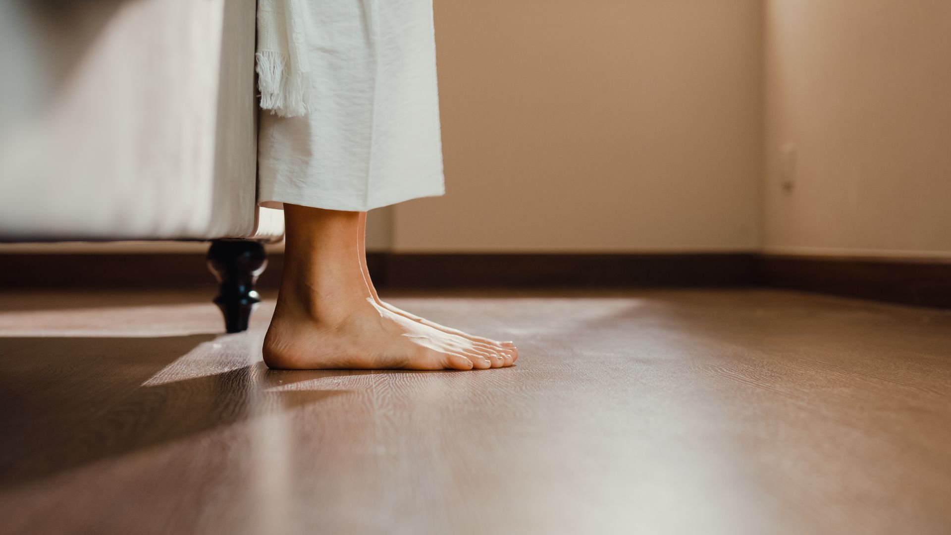 Des pieds nus près d'un lit sur un plancher en bois, laissant apparaître une robe de chambre blanche.