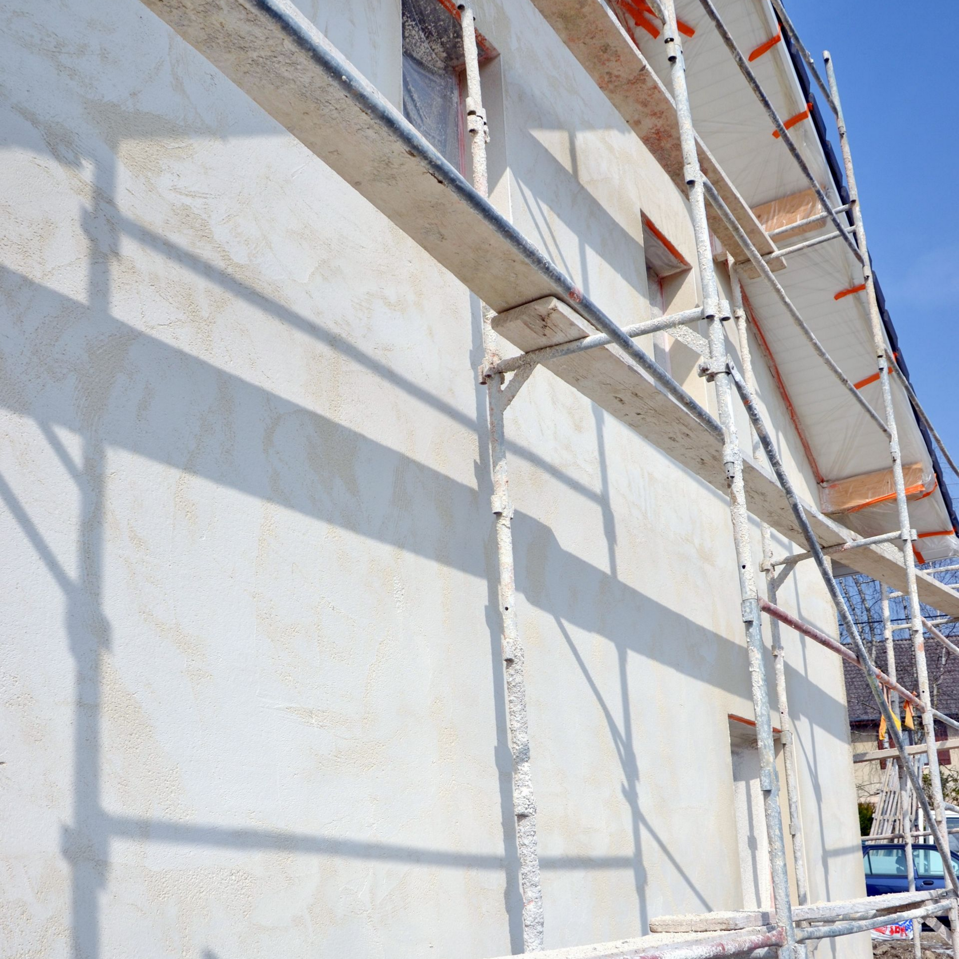 Échafaudages le long d'un mur blanc en construction, sur fond de ciel bleu et d'ombres.