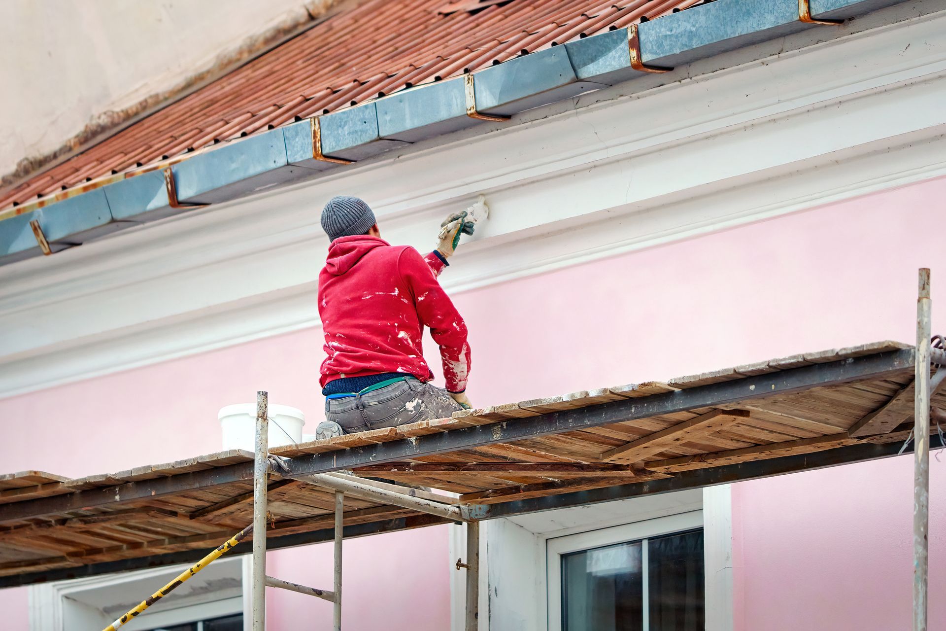 Un ouvrier en veste rouge peint un mur rose d'une maison depuis un échafaudage sous l'avant-toit.