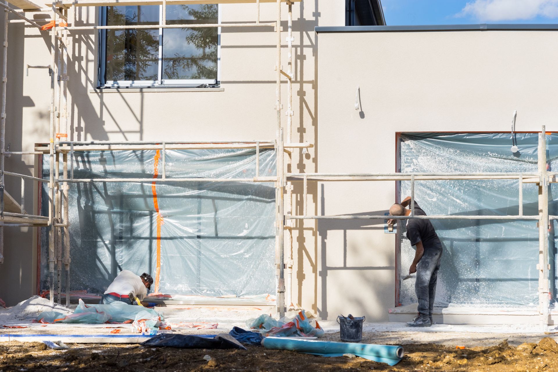 Des ouvriers, perchés sur des échafaudages, peignent la façade d'une maison. Des bâches bleues recouvrent les fenêtres et les matériaux situés en dessous.