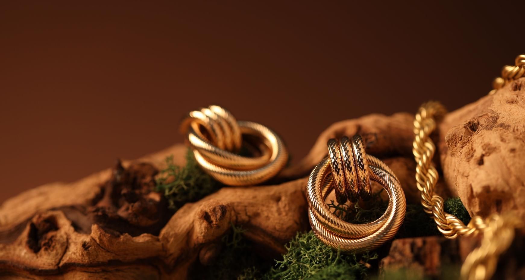 Anillos trenzados de oro y una cadena en una rama rústica con musgo, iluminada por una cálida luz marrón.