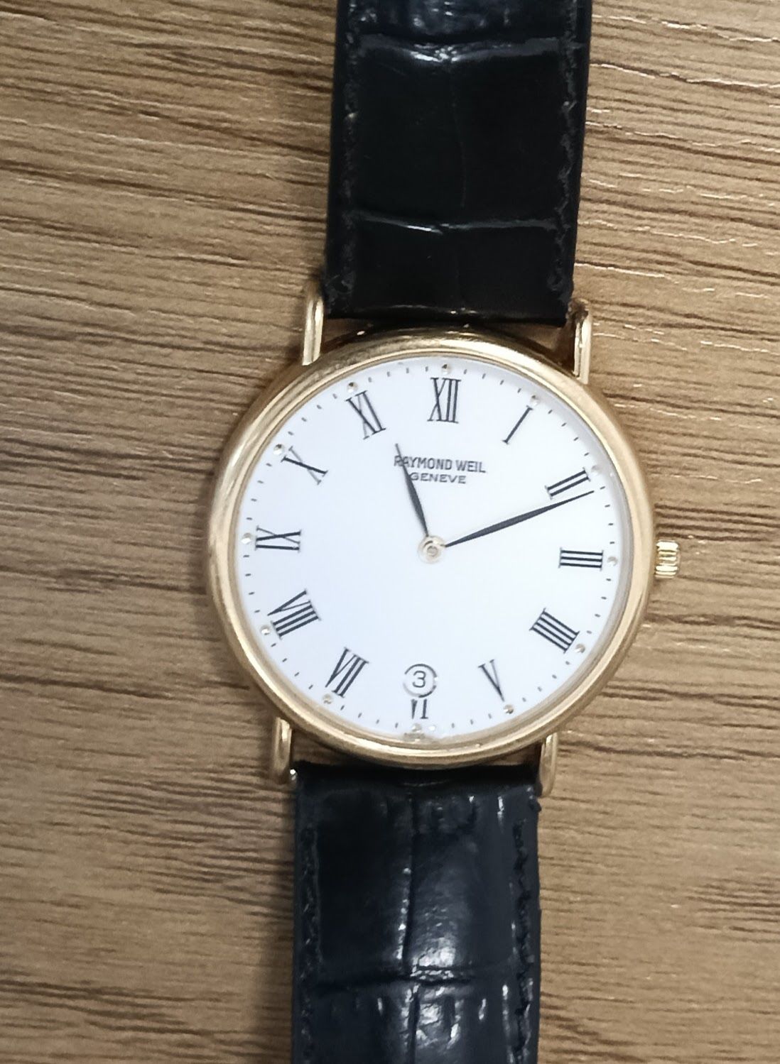 Reloj de pulsera dorado con esfera blanca con números romanos y correa de cuero negro sobre una superficie de madera.