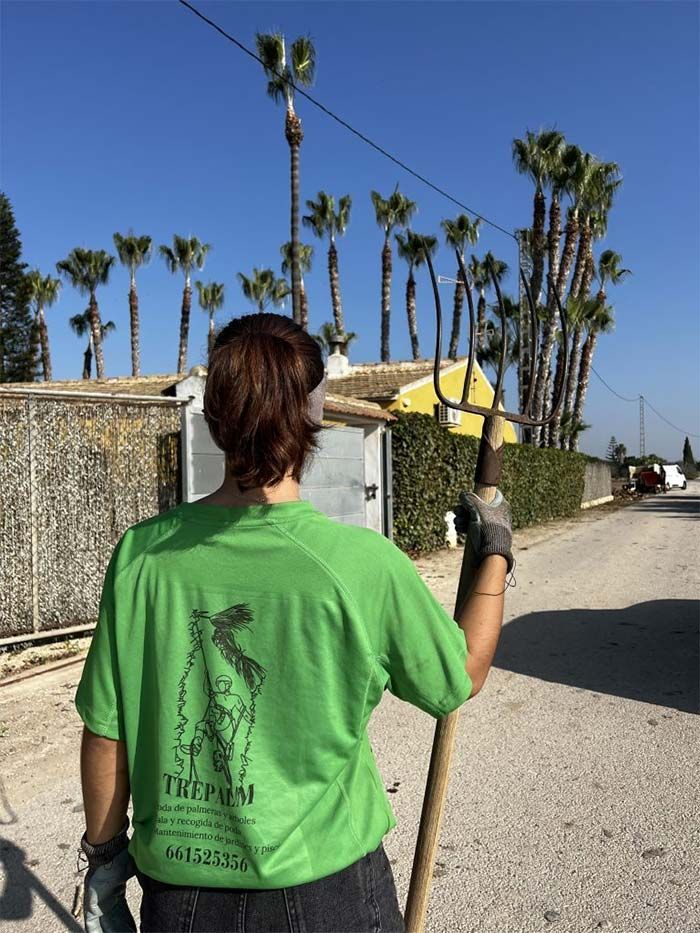 Una mujer con una camisa verde sostiene una pala frente a unas palmeras.