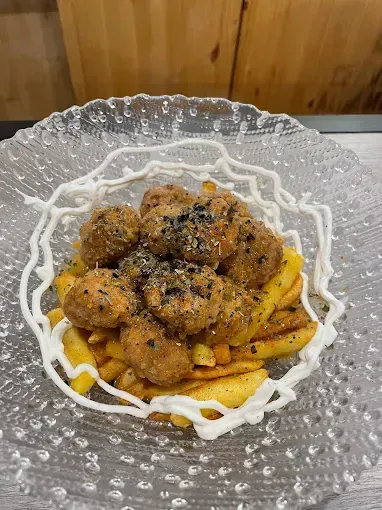 Trozos de pollo frito sobre patatas fritas, con salsa blanca decorativa, espolvoreados con semillas de sésamo negro, servidos en un plato de cristal.