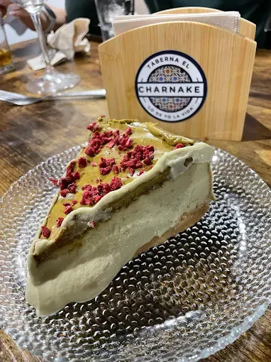 Rebanada de tarta de queso con pistachos y frambuesas secas, sobre un plato de cristal, cerca de un servilletero de madera.