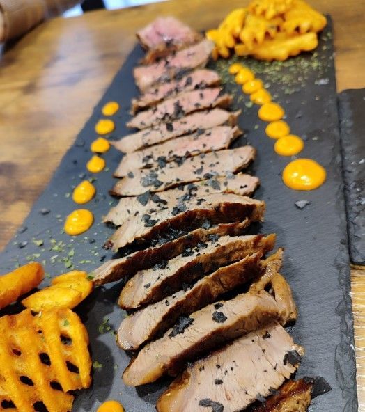 Filete en rodajas con patatas fritas enrejadas y salsa en un plato rectangular oscuro.