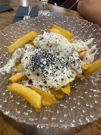 Papas fritas cubiertas con salsa blanca y motas negras sobre un plato de vidrio transparente.