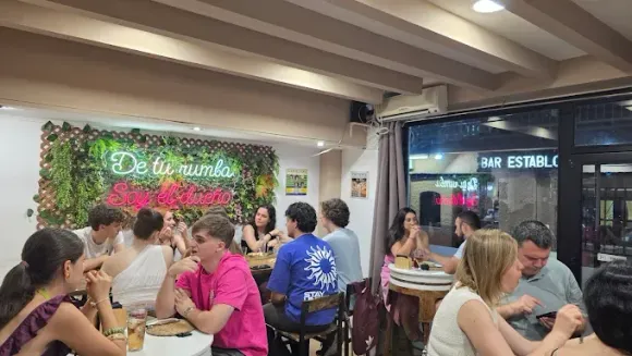 Personas sentadas en mesas en un restaurante con un muro vegetal y un letrero de neón. La luz brillante entra a raudales por la ventana exterior.