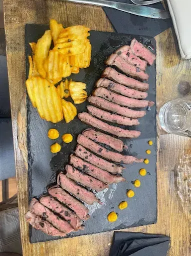 Filete en rodajas con patatas fritas y salsa en un plato de piedra negra, servido en una mesa de madera.