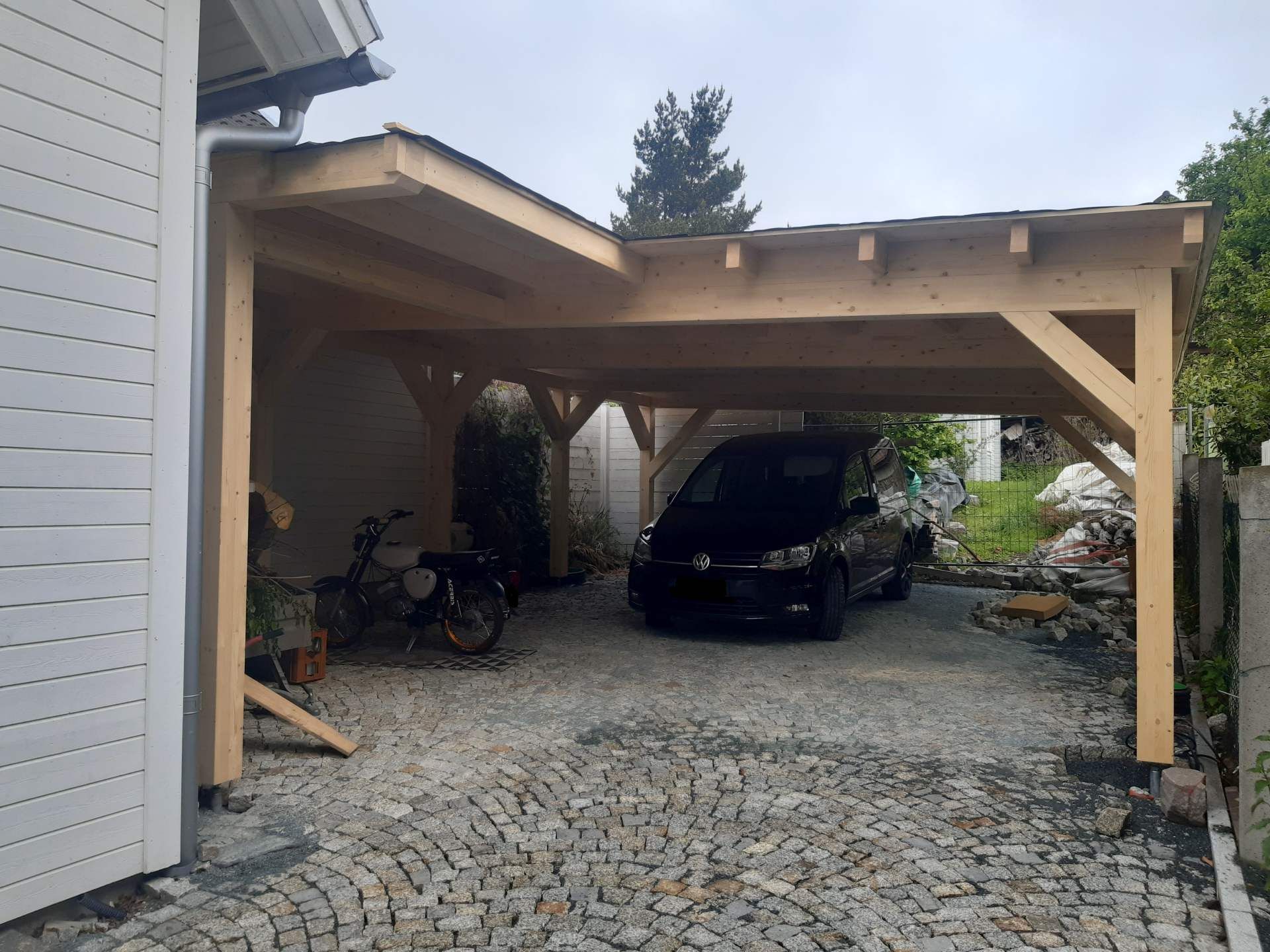 Ein hölzerner Carport, der an ein weißes Haus angebaut ist und ein schwarzes Auto und ein Fahrrad auf einer gesteinerten Auffahrt schützt.