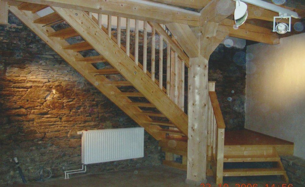Neben einer Steinmauer führt eine Holztreppe mit Balustern empor, an der sich ein vertikaler Stützpfeiler und ein weißer Heizkörper befinden.