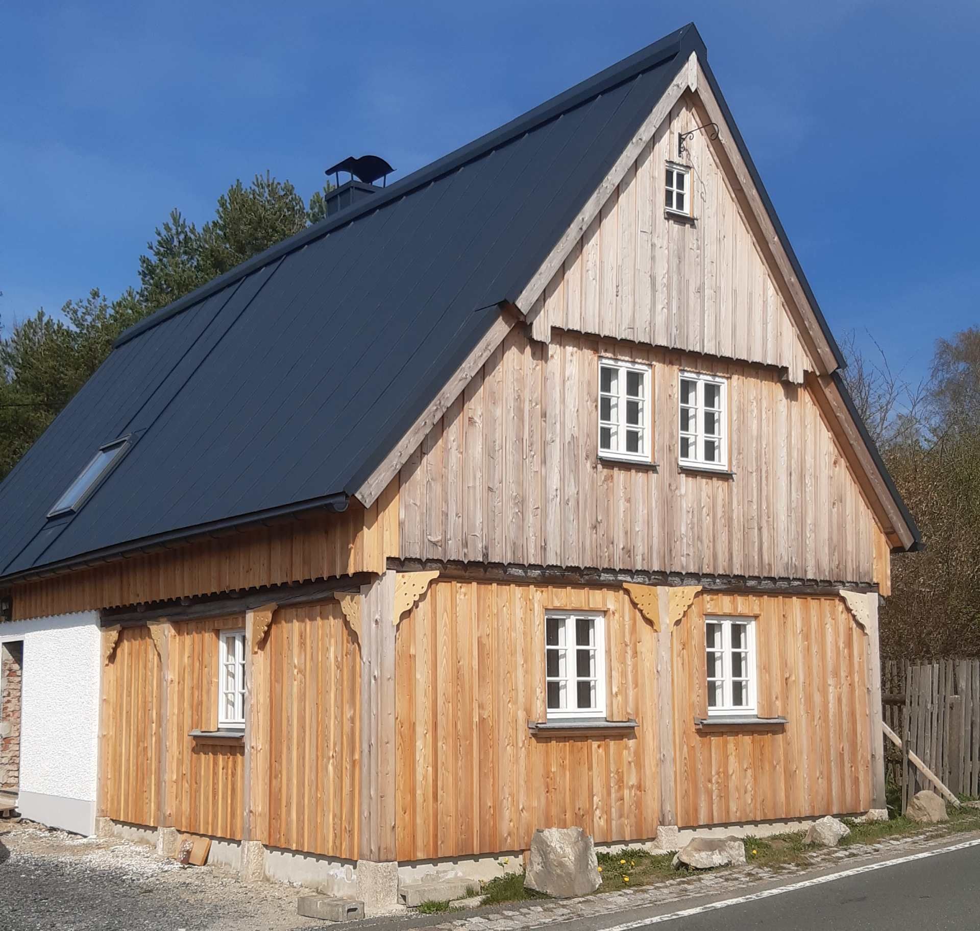 Ein zweistöckiges Haus mit naturbelassener Holzverkleidung, weiß gerahmten Fenstern und einem dunklen, schrägen Metalldach unter blauem Himmel.