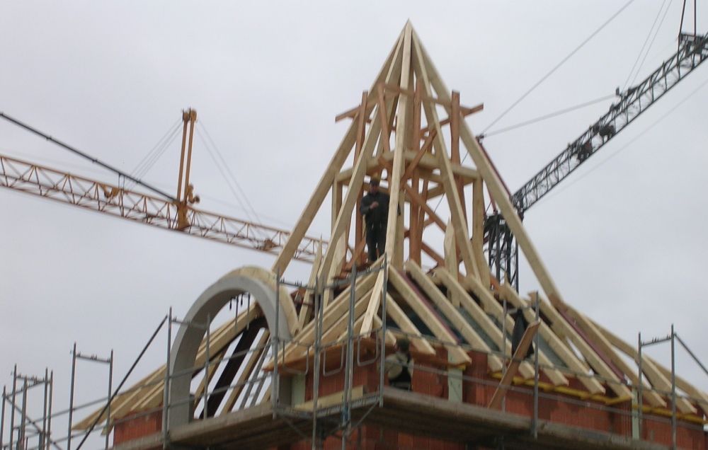 Ein Arbeiter montiert das Holzgerüst für ein steiles, spitzes Dach auf einem Backsteingebäude, während am Himmel Baukräne in den Himmel ragen.