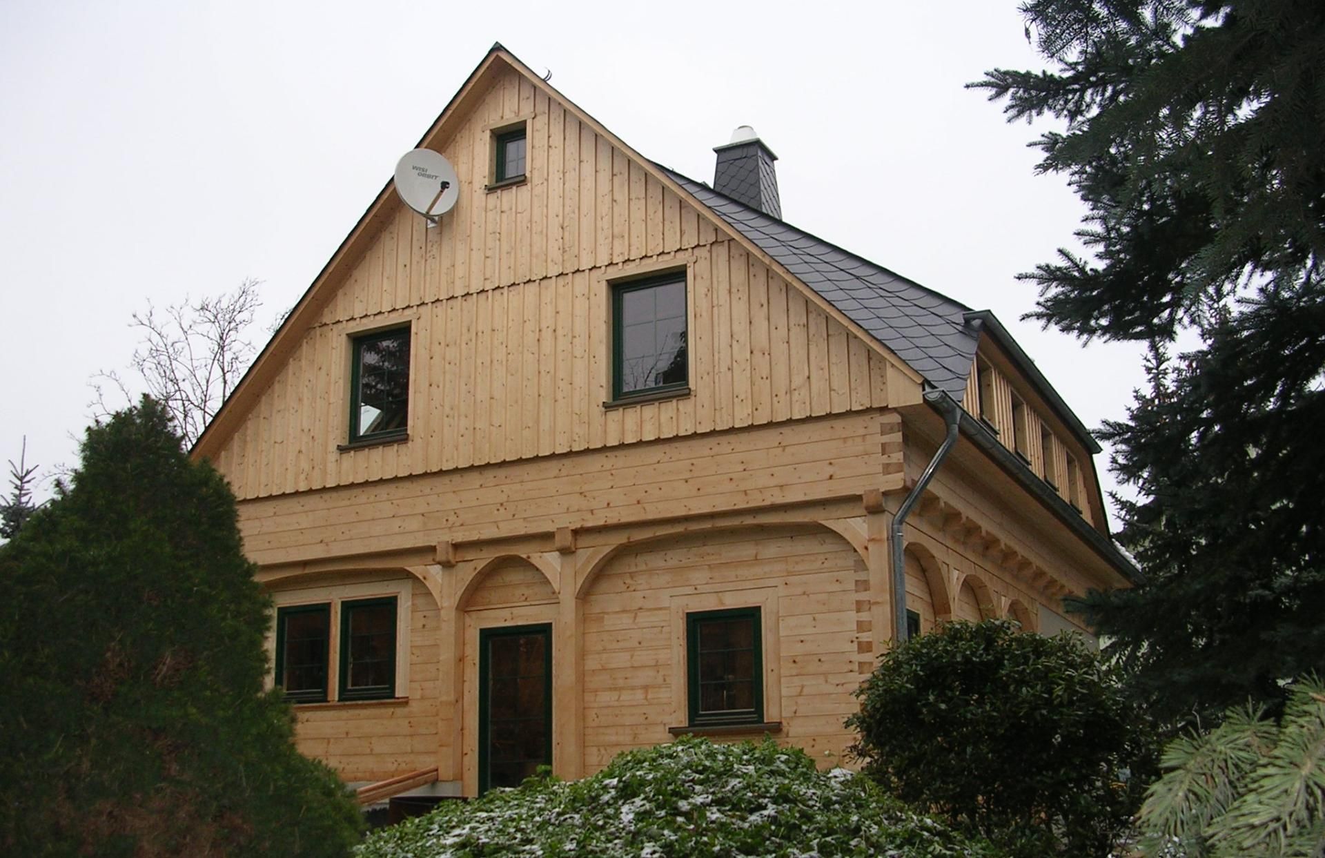 Ein zweistöckiges Blockhaus mit vertikaler Holzverkleidung am Giebel, gewölbten Verandastützen und einem grauen Schieferdach.