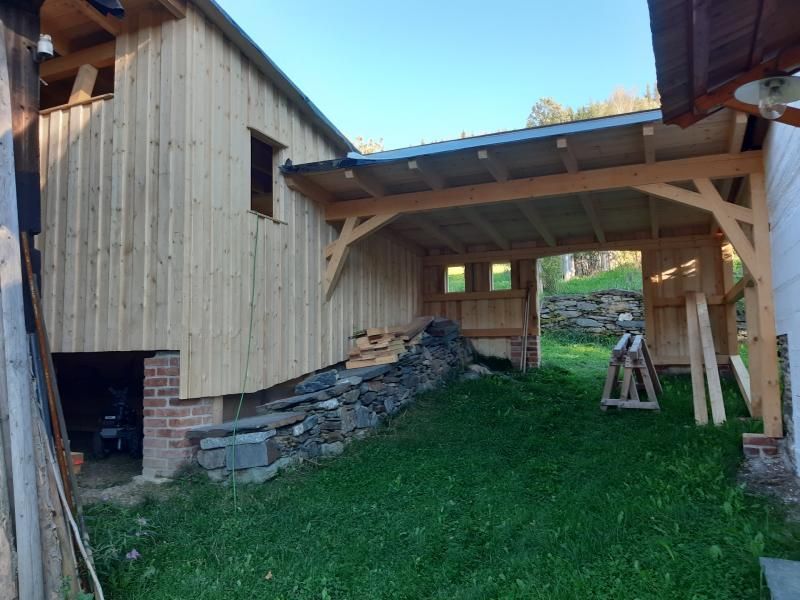 Eine hölzerne Scheunenkonstruktion mit überdachtem, offenem Außenbereich, Steinfundament und grasbewachsenem Vordergrund unter klarem, blauem Himmel.