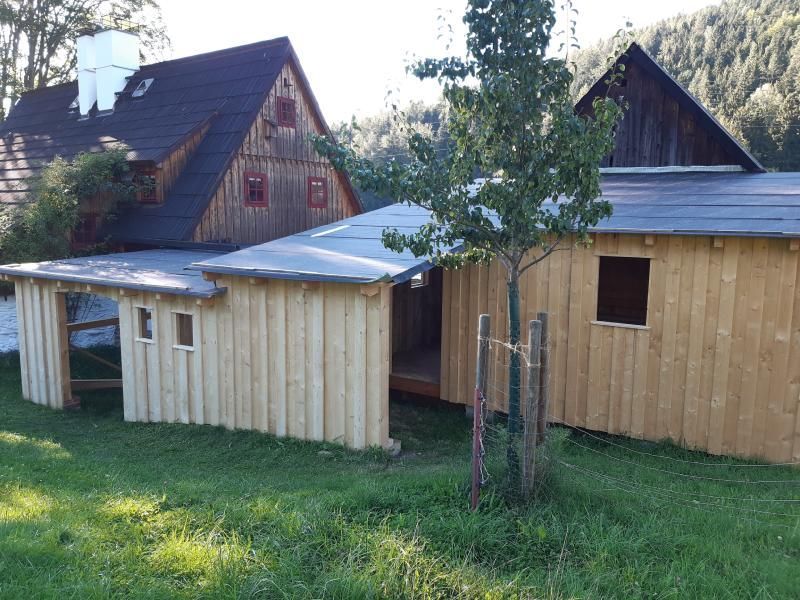 Ein neuer, hellholzfarbener Anbau, der an ein älteres, dunkelholzfarbenes Gebäude mit weißem Schornstein angebaut ist und an einem grasbewachsenen Hang liegt.