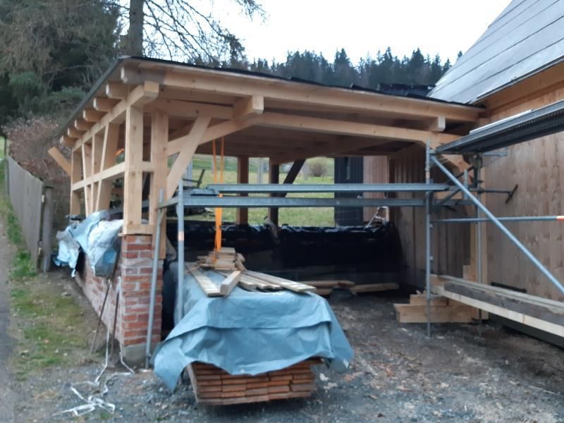 Ein im Bau befindlicher Carport aus Holz mit gemauerten Säulen, sichtbarem Fachwerk und vor Ort verwendeten Baumaterialien.