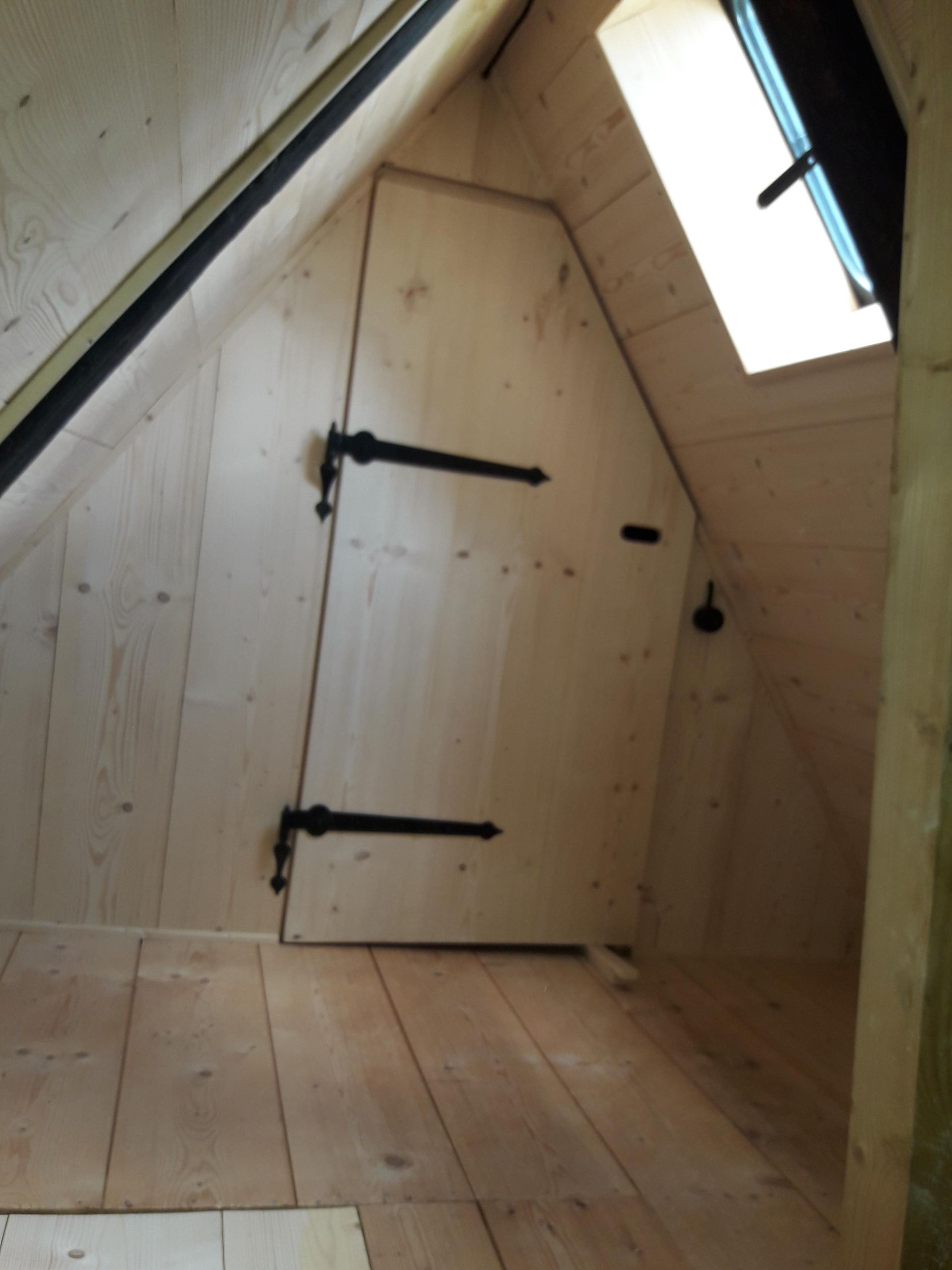 Ein Dachzimmer mit hellen Holzwänden und -boden, einer schrägen Decke, einem Oberlicht und einer kleinen, maßgefertigten Tür.