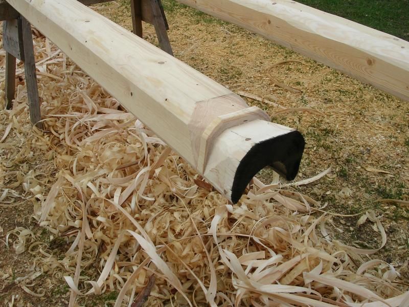 Ein Holzbalken mit einem gebogenen, dunkel lackierten Ende liegt im Freien auf einem Sägebock, umgeben von Holzspänen vom Schnitzen.