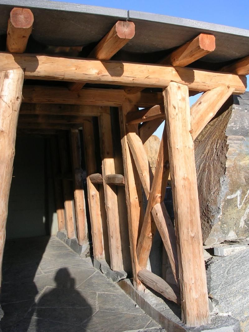 Eine Holzkonstruktion mit Stützbalken bildet einen überdachten Gehweg neben einer Steinmauer.