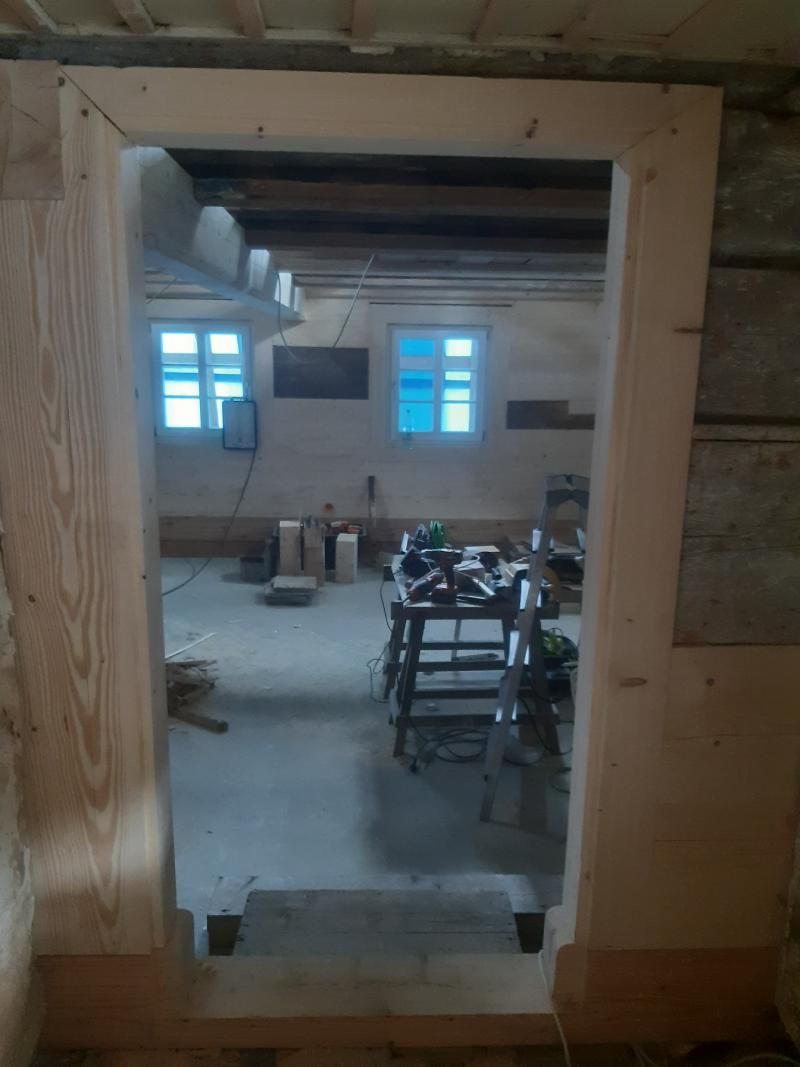 Ein hellfarbiger, im Bau befindlicher Holztürrahmen führt in einen Arbeitsbereich mit Fenstern und einer Leiter.