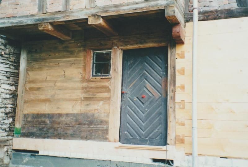 Die verwitterte Holzfassade des Gebäudes zeichnet sich durch eine dunkle Tür mit Zickzackmuster und ein kleines, quadratisches Fenster aus.