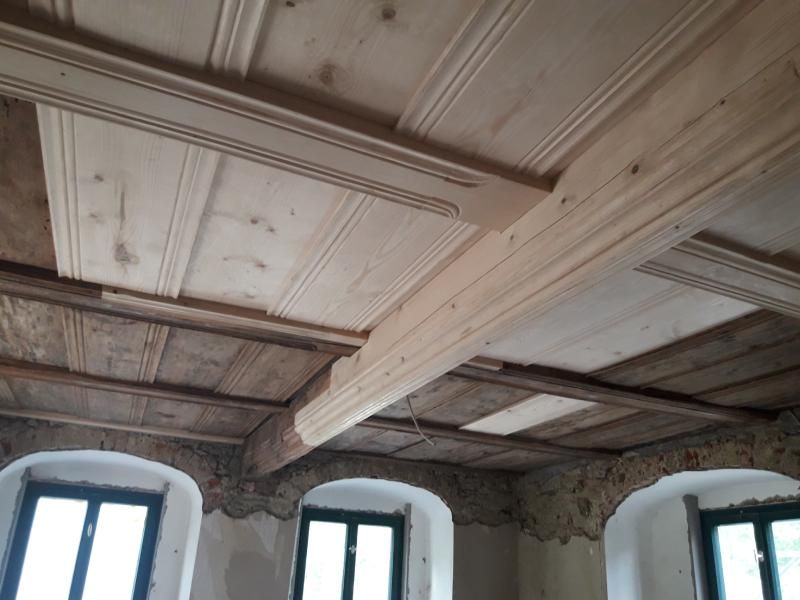 Teilweise restaurierte Holzdecke mit Zierbalken in einem Raum mit Bogenfensterrahmen und groben Steinwänden.